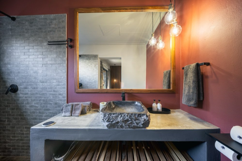 A carved stone basin on a concrete counter beneath a timber-framed mirror and hanging bulbs.
