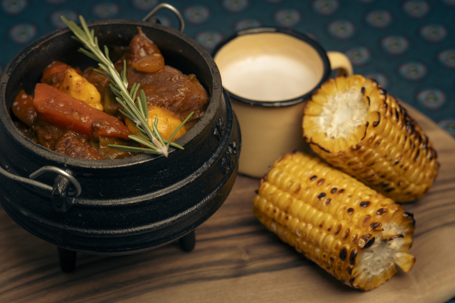 A cast-iron potjie of beef stew with rosemary on a wooden board, beside two cobs of charred corn and an enamel mug.