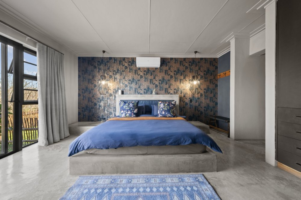 The Private Bush Cottage bedroom — a bed on a concrete plinth against patterned wallpaper, lit by two caged pendants.