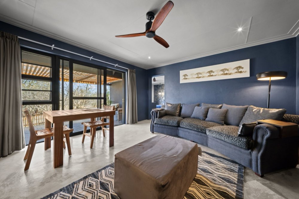 The Private Bush Cottage's own lounge — a long sofa against a deep blue wall, a timber dining table, and glass doors standing open onto the covered patio.