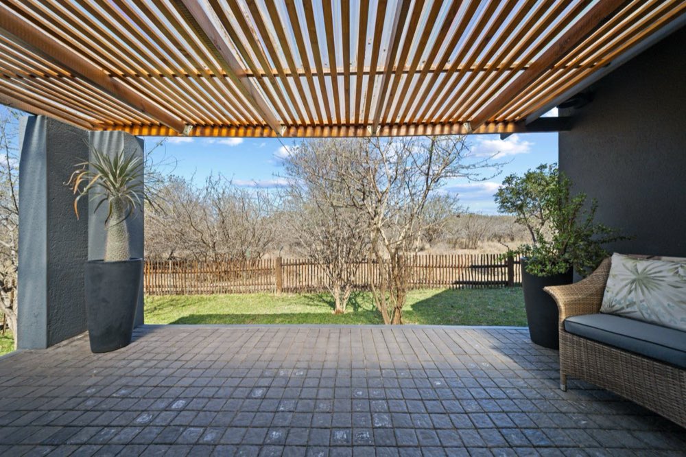 The Private Bush Cottage's covered patio under a timber pergola, looking past a reed fence to the bush and a low hill.
