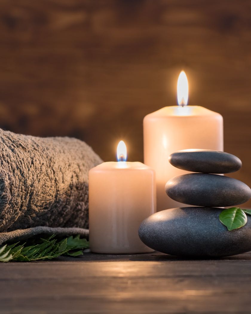 A rolled towel, two lit candles and a stack of smooth dark stones on a wooden surface.