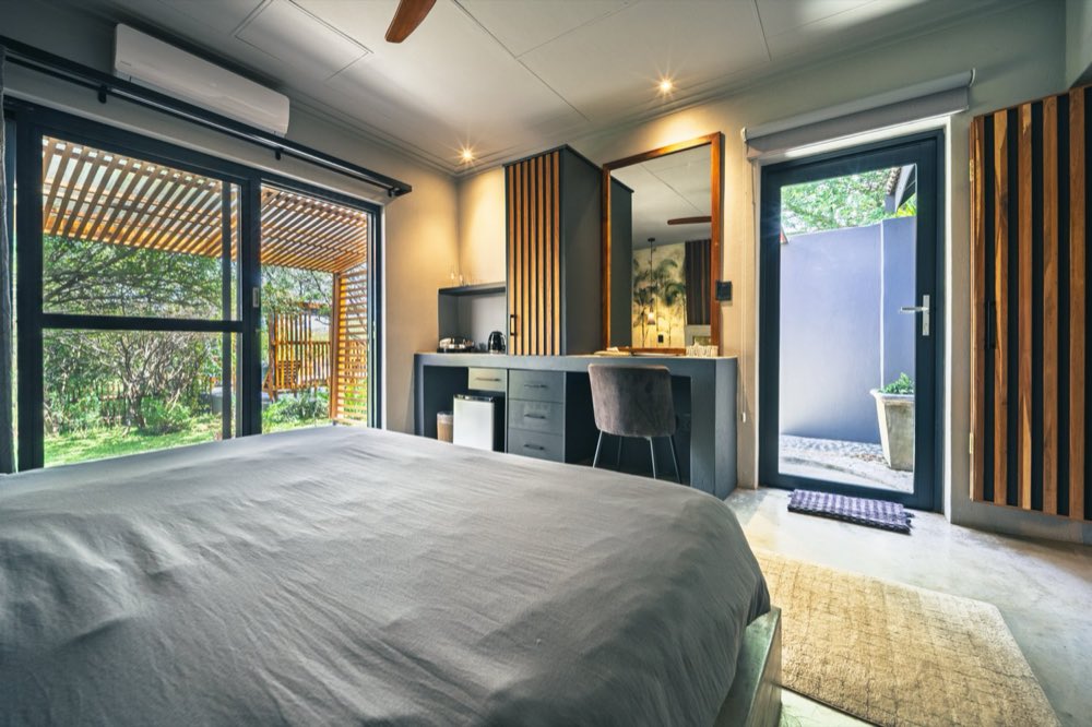 A room interior with doors open onto a shaded pergola, and a desk with a kettle, mirror and air conditioning above.